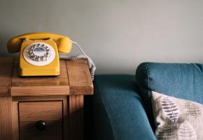 A yellow rotary dial phone on a side table, next to a blue sofa with a beige cushion on it.