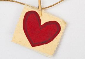 A red heart sewn onto cream fabric, hanging from a piece of string by a peg. 