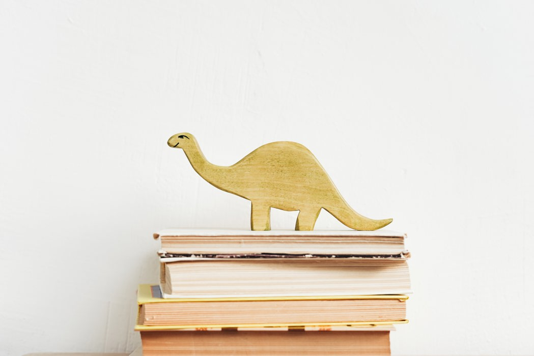 A wooden dinosaur toy on top of a stack of hardback books with their spines turned away from the camera