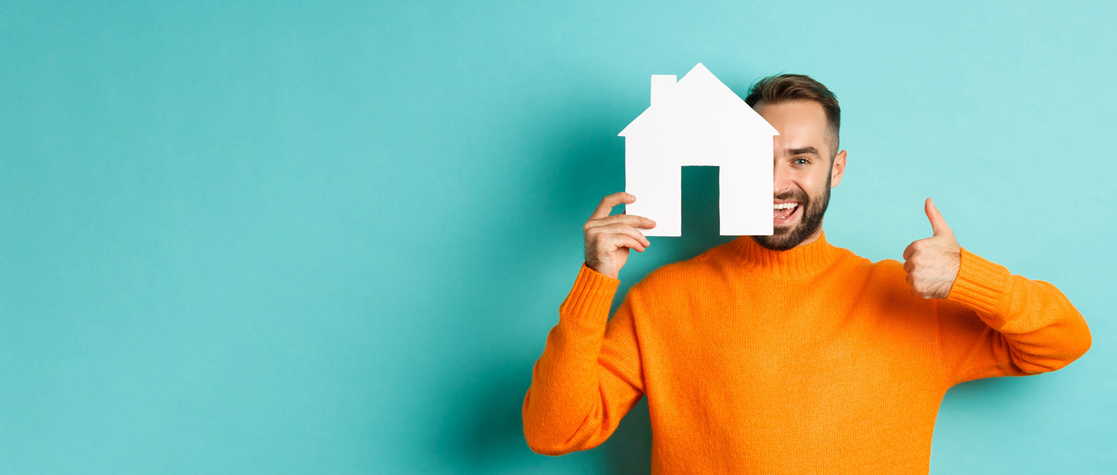 Photo of a smiling man in an orange sweater standing in front of a turquoise background. He is holding a white cut-out house shape half across his face and also giving us a thumbs-up sign