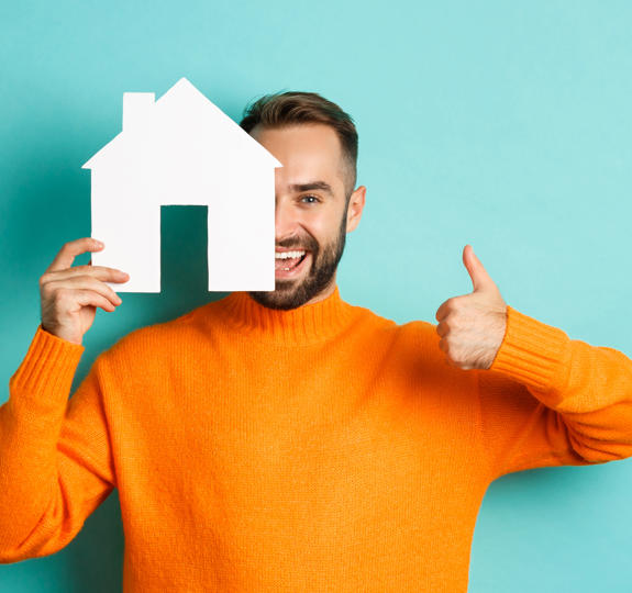 Photo of a smiling man in an orange sweater standing in front of a turquoise background. He is holding a white cut-out house shape half across his face and also giving us a thumbs-up sign