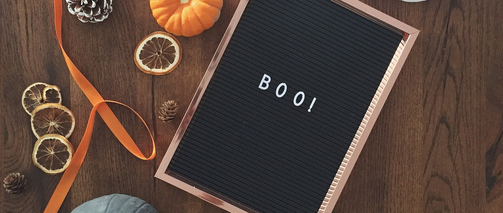 A pegboard reading "boo!" lying flat on a wooden table next to pumpkins of varying colours and sizes.