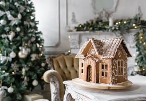 A gingerbread house in front of a Christmas tree and mantelpiece decorated in cream and gold.