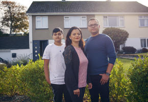 A teenage boy in a white t shirt, a woman wearing a black cardigan and a man in a blue jumper stood in front of a house.