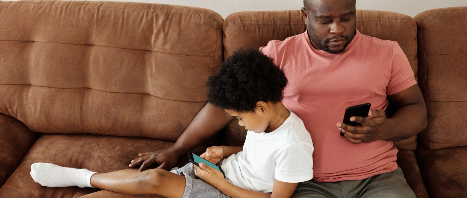 A child leaning against his father, both using mobile phones. They are sitting on a brown leather sofa.