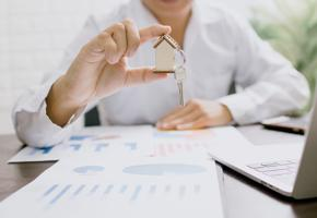 Photograph of someone holding a key with a house-shaped keyring over financial charts
