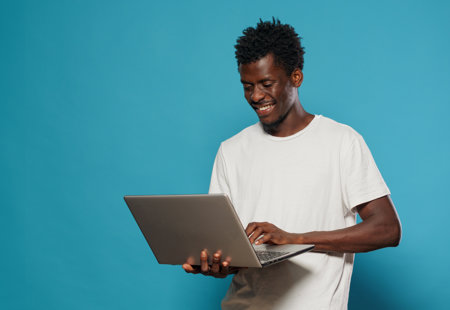 A young man wearing a white T-shirt holding a laptop. He is smiling and standing in front of a blue background.