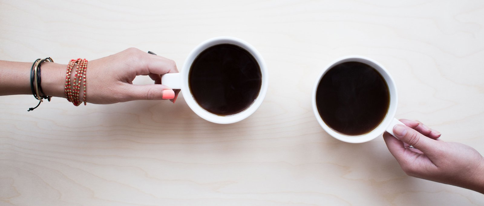 A shot from above of two mugs of black coffee, with a hand resting on each mug