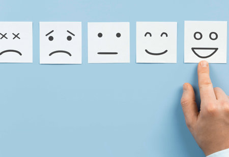 Photo of five faces drawn on white paper squared ranging from unhappy to very happy. A hand is pointing to the happy face. 