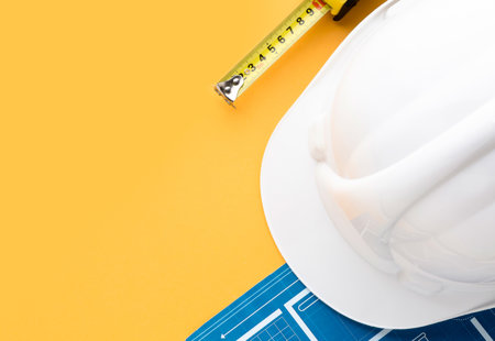 Photo of a white hard-hat, tape measure and some blueprints for a building on a yellow surface