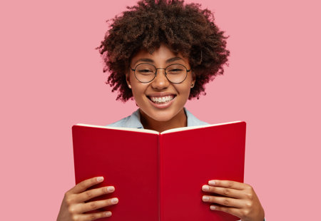Smiling woman posing against the pink wall while reading a book with red covers.